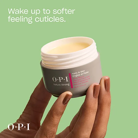 A hand with brown nail polish holds an open gray jar of OPI Nature Strong cuticle balm with yellowish contents, displaying text that reads Wake up to softer feeling cuticles, OPI, Nails & Skin, Ongles et Peau, To the Rescue, Overnight Cuticle Balm, Baume Reparateur de Nuit pour Cuticules, 20 mL / 0.7 fl. oz.